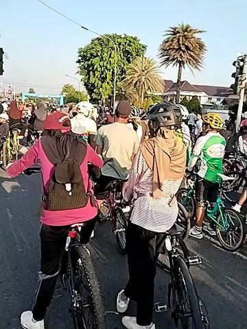 The video opens with a wide shot of a busy street, where numerous individuals, mostly adults, are seen gathered with their bicycles. A few people are still, while others are slowly moving forward, navigating through the crowded area. Many of them are wearing helmets, and some are wearing long-sleeved shirts, possibly to protect themselves from the sun. The sun is clearly visible in the sky, casting long shadows on the ground. In the background, there are buildings and trees, including palm trees, suggesting a warm climate. A large street light with multiple arms is also visible in the frame. The video ends with the crowd continuing to move along the street.
