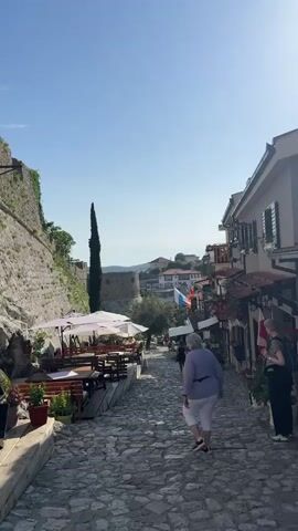 The video shows a cobblestone street in a European town, with a stone wall on the left and buildings on the right. A woman in white pants and a purple shirt is walking down the street, away from the camera. On the left, there's an outdoor seating area with tables and umbrellas. In the distance, a fortress or castle is visible on a hill. Flags are displayed near the buildings on the right, and a person with a backpack is looking into a shop. The sky is clear and blue.