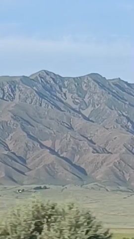 A wide shot shows a vast, mountainous landscape. The mountains, with their rugged peaks and slopes, are a dusty brown and gray, with shadows defining their contours. In the foreground, a rolling green plain stretches out, dotted with sparse vegetation and a few buildings in the distance. The sky above is a clear, pale blue. Electrical wires are visible, strung across the frame in the upper portion, suggesting human presence and infrastructure in the otherwise wild and natural setting.