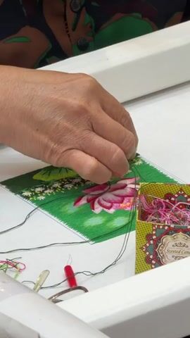 A close-up shows a person's hands embroidering a design. The design features a pink flower (possibly a lotus) surrounded by green leaves and small white flowers, embroidered on a white fabric stretched within a rectangular frame. A small, colorful box labeled "Scented Candle" sits to the right of the embroidery. The box contains spools of brightly colored thread. A pair of small scissors and a red needle threader are also visible on the table. The person is using dark green thread to add details to the embroidery, manipulating the fabric and thread with their fingers. The background is a white surface, possibly a table. The person wears a shirt with a dark, patterned design.