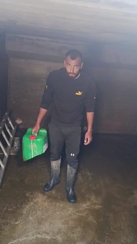 A bearded man stands indoors holding a green container. He wears a black Puma shirt, black Under Armour pants, and black rubber boots. The room is small with a low concrete ceiling.  The floor is wet and partially covered in dark sediment. A ladder stands on its side in the background.