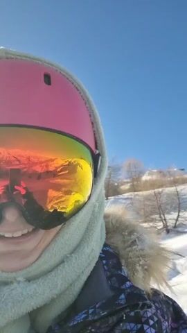 A woman wearing a pink helmet with mirrored goggles, a light green scarf around her head, and a jacket with a geometric pattern on it is filming herself in the snow on a sunny day. The reflection in her goggles reveals mountains in the distance. The camera then pans to the side, revealing a snowboard in the snow next to her and a man in snowboarding gear standing on a snowboard. The landscape behind them features snow-covered hills with trees and a ski lift in the distance.
