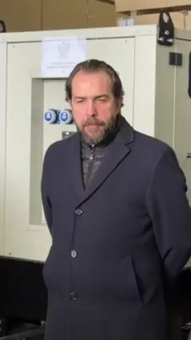 A medium shot shows a man in a dark coat standing in front of an electrical generator. The man has dark hair and a beard, and he is looking directly at the camera with a serious expression. He is standing with his hands behind his back. The generator behind him is a large, beige-colored box with a sign in Polish on the front. The background is a neutral indoor space. The camera is steady. The man gestures with his right hand.