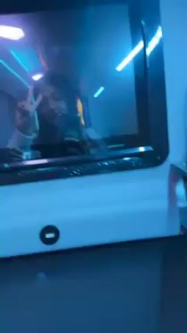 The video shows a woman taking a selfie in a mirror, making a peace sign and then a thumbs up. The mirror is part of a white structure that has blue neon lights reflected in it. There is a USB port on the white structure below the mirror. The environment seems to be a vehicle, potentially a bus, with blue lighting.