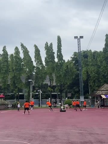This video shows a group of people playing a sport on a red court. The players are wearing orange shirts and appear to be engaged in a game of sepak takraw, a sport native to Southeast Asia. Several trees line the background of the court, and a building with a brown roof is visible to the right. The sky is overcast, suggesting cloudy weather. A tall light pole stands in the center of the background. The players are spread across the court, with some actively participating in the game and others observing.