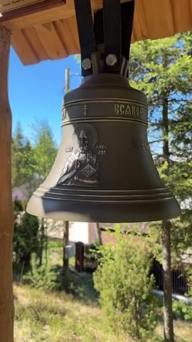 Here's a description of the video:

The video shows a row of Orthodox church bells hanging from a wooden structure outdoors. The bells are bronze-colored and have inscriptions in Cyrillic script. One bell features a depiction of a saint. A hand reaches into the frame to gently strike the bells. The background shows trees and a bright blue sky, suggesting a clear, sunny day in a natural setting. The video is shot from a low angle, looking up at the bells.