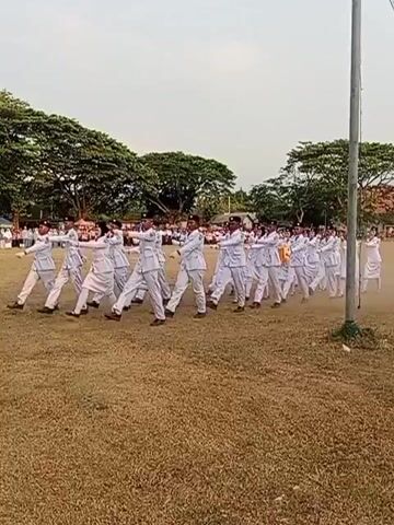 A group of young people in white uniforms are marching on a grassy field. They appear to be participating in a drill or parade. The field is mostly dry grass with some trees in the background. The sky is overcast. There is a tall pole on the right side of the frame. The people are marching in formation and appear to be in sync.