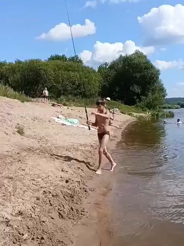 A young person wearing swim trunks stands on a sandy bank next to a body of water, holding a fishing rod. The fishing line extends into the water. There are other people in the background on the beach, some of whom appear to be swimming or relaxing. Trees and foliage line the far bank of the water. The sky is blue with scattered clouds. The scene appears to be a warm, sunny day at a recreational area.