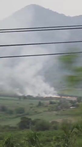 The video depicts a fire burning in a field, with a backdrop of hills and trees under an overcast sky. The scene is partially obscured by overhead power lines. The perspective appears to be from a moving vehicle, as the view quickly transitions to the interior of the vehicle and then to a blank, light-colored surface.