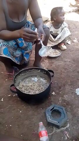 A woman is squatting outside near a young child, who also squats. The woman is holding a spoon and a bowl and seems to be preparing a meal from a large pot on the ground in front of her. A man peers at the woman, and the camera turns to show two bare-chested men in the background squatting near the pot. One man is wearing a yellow shirt. The environment is an unpaved outdoor space.