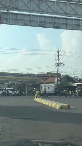 A wide shot captures a street scene, likely an intersection, on a bright, clear day. In the foreground, the edge of a car's dashboard is visible. The road surface is asphalt, and a raised speed bump with yellow and black markings runs across the frame. A row of motorcycles and a few cars are present on the road. A small convenience store, possibly an Indomaret, with a blue and yellow awning, is visible in the background. Several people, some wearing reflective vests, appear to be directing traffic or working. Utility poles and power lines crisscross the sky. The overall impression is of an active urban environment.