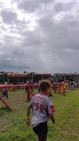 A video shows an outdoor historical reenactment event with people dressed in medieval-style clothing. In the foreground, a young male wearing a grey t-shirt with the number 159 on the back is walking away from the camera, towards the reenactment area. The reenactment area features a fenced-in grassy field with spectators seated in bleachers in the background. Several individuals in the field are dressed in yellow and red tunics and appear to be involved in a staged combat or demonstration, possibly with spears or other weapons. The sky is cloudy.