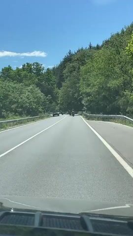 A dash camera perspective shows a two-lane road winding through a forest. The road has a white solid line on the right and a broken white line to the left. A silver car is ahead and to the left, and a group of motorcycles are ahead in the right lane. The motorcycles are moving forward, and the car is also moving forward. The trees are green and lush, indicating a warm season. The sky is blue with some white clouds. The road appears to be well-maintained.