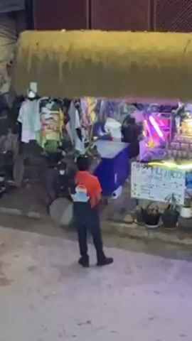 A person in an orange shirt and black pants and shoes is standing on a dirt road facing a colorful vendor stand. The vendor stand has a thatched roof with clothing and other merchandise hanging from it. Behind the vendor stand, there is a building with a sign that reads "SWAN RAY." The person raises their arms and moves them around as if dancing or exercising.