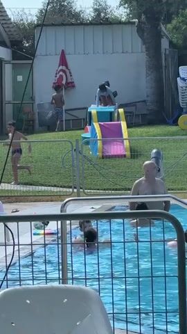 A group of people are at a swimming pool on a sunny day. In the foreground, partially obscured by a white plastic chair and a metal fence, is a bright blue swimming pool filled with clear water. Several children are in the pool, with one appearing to be swimming and another partially submerged. An adult male with a shaved head is standing in the pool. In the background, beyond the pool fence, a grassy area is visible. A colorful plastic slide with yellow, blue, and pink sections is set up on the grass. Two young boys are near the slide, one in a striped swimsuit standing and holding something, and another boy in a blue top and dark shorts. A young child in a light blue top and dark bottoms is on top of the slide. A red umbrella with a white logo is propped against a white building. A palm tree stands to the right of the building, and other playground equipment is visible. The overall scene suggests a leisurely day at a recreational facility.