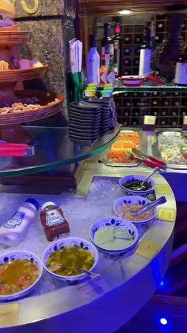The video captures a close-up, panning shot of a buffet display in what appears to be a restaurant. The camera begins by focusing on a three-tiered wooden tray laden with various food items like bread and nuts, positioned on a glass surface. Below the glass, several bottles of condiments such as ketchup, mustard, and mayonnaise are nestled in ice, alongside numerous small white bowls filled with different sauces or dips. To the left of the frame, a pair of metal tongs with red handles rests on a white plate. As the camera pans upwards and to the left, it reveals a wall densely decorated with an eclectic assortment of decorative plates and other ornamental objects, including a white rocking horse figurine, a small guitar, and various colorful dishes of different sizes and designs. Blue lighting illuminates parts of the wall and the buffet area, adding to the ambiance. A small bottle of hand sanitizer is also visible on a wooden surface near the wall decorations. The video ends with a view of the intricate wall decor.