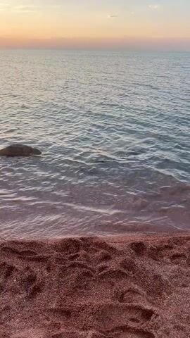 The video shows a sunset over a body of water. The foreground is a sandy beach with footprints. In the middle ground, the water is calm with small waves. There is a large rock partially submerged in the water. In the distance, two people are on a paddleboard. The sky is a gradient of orange, pink, and blue with scattered clouds.
