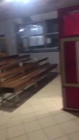 The video shows the interior of a UBA (United Bank for Africa) branch. Initially, a red booth with U-Mobile signage is visible. The camera pans to reveal an empty waiting area with rows of wooden benches, some chairs, and advertising displays. The space is illuminated by overhead fluorescent lighting, and windows are visible along one wall. The camera moves slightly, showing more of the seating area. The environment appears clean but deserted.