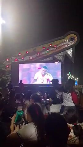 Here is a description of the video:
The video shows a crowd of people seated outside at night, watching a large screen showing video clips of sporting events. The screen is set up on a stage-like structure. The stage has a large alligator head shaped structure above it, decorated with lights. People are sitting in chairs, some are eating and drinking. Some are looking at their phones. There's a street lamp to the left of the screen, creating a bright spot in the frame. In the background, you can see street lights and lit-up Christmas tree shaped decoration. The camera is positioned behind the crowd, capturing their reactions to the screen.