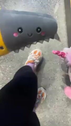 Here is a description of the video:
The video shows a person holding a plush toy hand saw. The saw toy is yellow and gray, with a shark-like face on the blade part. The person holding the toy is wearing black pants. Beside them is a pink unicorn plush toy. The video then shows the person holding the saw and unicorn toys above their head, revealing a light brown wood ceiling with track lighting. The person is wearing a black Adidas sweatshirt and a white headband.