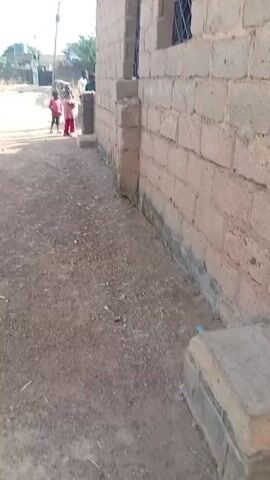 The video shows a small, unpaved path running alongside a brick building. There are two small children on the path, one wearing a red shirt and the other wearing a pink shirt. Further down the path, near the building, are several other people standing near the corner of the building. The building has two barred windows visible in the frame. The path is made of packed dirt and small pebbles. In the distance, there are houses and a water tower visible. The sky is bright and sunny. There is a concrete block in the foreground of the video. A goat passes the group near the building.