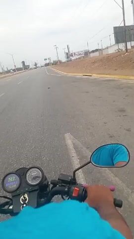 A first-person view from a motorcycle traveling down a paved road. The speedometer shows the motorcycle's speed. The rider wears a blue shirt and has their right hand on the handlebar, which has a pink grip. The road has a solid white line dividing lanes. There are other vehicles visible in the distance. Power lines run along the right side of the road, and a large sign for "CONEL" is also visible on the right. The weather appears overcast.