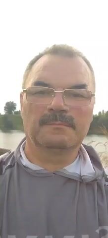 A close-up, head-and-shoulders shot shows a middle-aged man with a mustache and glasses looking directly at the camera. He is wearing a gray hooded sweatshirt over a white shirt. The background is a blurred outdoor scene with a body of water and trees under a bright sky. The video is very short and shows no action.