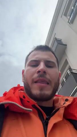 A man wearing an orange work jacket is looking at the camera and says “Fuck you Nathan”. He has brown hair and a short brown beard. He is standing outside on a cloudy day. There is a building behind him with a sign in French.