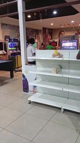 The video shows an indoor scene which appears to be an arcade or entertainment venue. In the foreground, there are several empty white shelves on a metal rack. To the left, there is a pool table with a dark surface. Behind the shelves, a person wearing a white shirt walks away from the camera. Further in the background, there are arcade game machines, including one labeled "Mortal Kombat." Several people are visible in the background, some appearing to be interacting with the games or with each other. The environment is brightly lit, and the floor is tiled. The overall impression is a commercial space dedicated to entertainment.