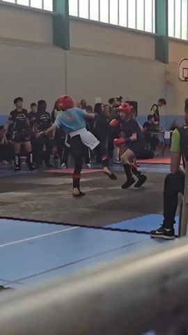 Here's a description of the video:

The video shows two young individuals engaged in a kickboxing match. They are both wearing protective headgear and gloves. They are positioned on a black mat, likely the fighting area, surrounded by onlookers. The environment is a gymnasium with blue floors. In the background, there is a basketball hoop and various people watching the match, including coaches and competitors. A man is sitting on a chair in the foreground, observing the fight. The young fighters are exchanging blows.