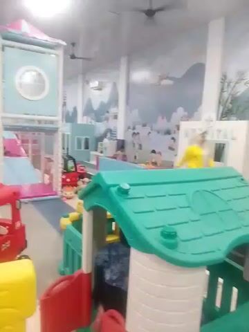The video shows a bright, indoor playground designed for children. The scene is filled with colorful play structures, including a multi-level soft play area with slides and ball pits, a small red toy truck, and a yellow crocodile-shaped seesaw. The walls are decorated with a whimsical mural depicting a village scene with children playing and trees.

Several people are present in the playground, including adults and children engaged in play. One adult, wearing a hijab, is seated on a bench in the background, observing the children. Another adult is partially visible on the left side of the frame. Children can be seen in the ball pit and on the play structures.

The overall atmosphere appears to be cheerful and lively, typical of a children's play area. The lighting suggests it is daytime, but the indoor setting makes it independent of weather conditions. The presence of specific toys and the mural's design point towards a family-friendly entertainment venue. While the exact time of day isn't discernible, the clean and well-lit environment suggests it's open for business. No specific events or celebrations are indicated. The location context "Rawang Itiek, Indonesia" is provided, identifying the general area where this playground is situated. There is some text visible on the mural, but it's too small and blurry to read. The word "HOSPITAL" is clearly visible on a small white building structure in the background, which appears to be part of the play area.