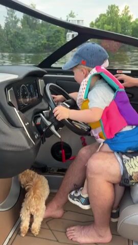 A toddler wearing a blue baseball cap and a colorful life vest sits on an adult's lap, holding the steering wheel of a boat. The adult, wearing sunglasses and a black baseball cap, sits beside the toddler, with one hand on the steering wheel and the other resting on the toddler's shoulder. A small, light-brown dog stands near the adult's feet on the boat's floor. The boat is on the water, with trees and a modern house visible in the background on the shore. The dashboard of the boat displays screens and controls. The video captures the toddler seemingly operating the boat, with the adult supervising.