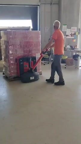 A man wearing an orange t-shirt and grey pants is operating a red and black pallet jack. He is moving a large pallet stacked high with cases of red and white cans towards an open loading dock door. The pallet is wrapped in plastic. The man is facing away from the camera and appears to be pushing the pallet into the loading dock. The floor is a light grey concrete. In the background, there are office chairs, a desk with a computer monitor, and some safety vests hanging on a wall. The lighting suggests it is daytime.