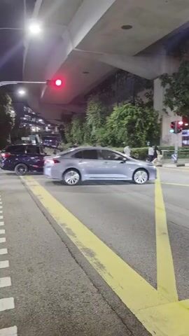 A dark blue SUV is turning left across a road at night. A silver sedan is stopped, waiting for the SUV to pass. In the background, a double-decker bus is stopped at a traffic light. The scene is under an overpass, with trees and streetlights visible. There are yellow road markings on the asphalt.
