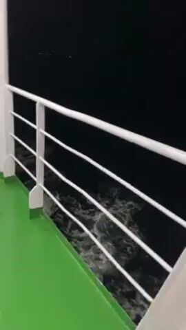 The video depicts a view from the deck of a moving vessel at night. The deck is painted green and bordered by a white railing. A red and blue life preserver hangs from the railing. The sea is dark, but the vessel's wake is visible as white foamy water churning against the blackness.  Small lights can be seen in the distance. The camera appears to be stationary, focused on the wake.