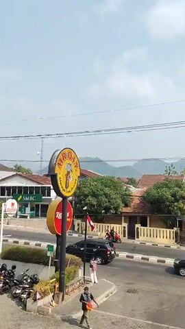 The video begins with a street scene, showcasing a variety of vehicles including cars and motorcycles. The camera pans across a street lined with buildings, some of which appear to be shops or restaurants, evidenced by signage like "MADIN". There are people on the street, walking and riding motorcycles. The environment appears to be a town or city, with houses and trees visible in the background, and rolling hills in the distance under a bright, cloudy sky. The video appears to capture a moment in time of everyday life on a busy street.