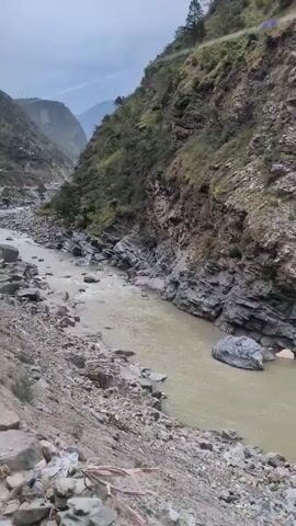 The video shows a muddy river flowing through a rocky canyon. The riverbanks are steep and covered with loose rocks and vegetation. The water appears to be fast-moving. The sky is overcast and the light is dim, suggesting it is either early morning, late evening, or a cloudy day. There are no people or animals visible in the video.