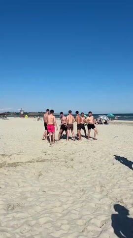 A group of young men are gathered on a sandy beach under a clear blue sky. They are all wearing swim trunks, and some are shirtless. The men appear to be interacting, possibly playing a game or engaging in conversation. In the background, the ocean is visible, with gentle waves and the horizon line. There are a few scattered beach umbrellas and people in the distance, suggesting it's a public beach on a sunny day. The sand is light in color and appears dry. The overall atmosphere seems relaxed and recreational.