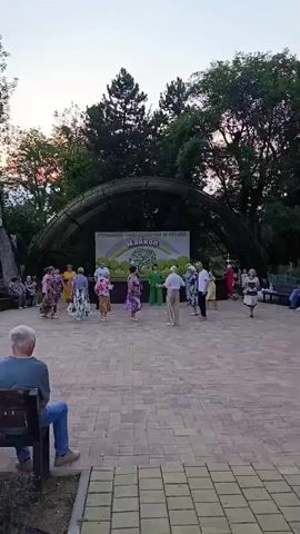 A group of people are dancing in an outdoor amphitheater. There are trees and a clear sky in the background. Some people are seated on a bench watching the dancers.
