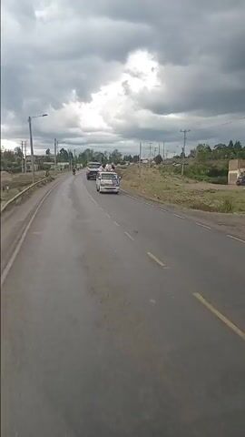 The video captures a forward-facing view from a moving vehicle on a two-lane paved road under an overcast sky. The weather appears cloudy, with some dark clouds overhead. The road itself shows signs of wear and tear, with some wet patches, possibly from recent rain.

**Subjects and Activity:**
The road is moderately busy with various types of traffic. Vehicles observed include:
- Several cars (white, blue, black, and a small green minivan).
- A large white dump truck.
- Multiple motorcycles (boda-bodas), some carrying passengers, moving in both directions.
- At least two matatus (Kenyan public transport minibuses), one with luggage on its roof, and another white and yellow one parked on the side.
Pedestrians are visible walking along the roadside or standing near parked vehicles. At one point, two people are seen with a motorcycle by the roadside, and a cow is also grazing near the road shoulder.

**Scene and Setting:**
The setting appears to be a semi-urban or rural area along a main road. On both sides of the road, there are dirt shoulders, some sparse vegetation, and scattered buildings or structures in the distance. Utility poles with power lines run parallel to the road. Streetlights are also present.

**Notable Details:**
- The footage is captured during the daytime.
- The overcast sky suggests dull lighting conditions.
- The road has clear white and yellow lane markings.
- The presence of matatus and numerous motorcycles indicates a typical Kenyan road scene.

**Visible Text:**
- A billboard on the right side of the road prominently displays the word "SAVANNAH."
- The white and yellow matatu has "MADARAKA" written on its side.