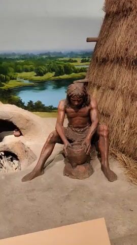 The video shows a diorama depicting prehistoric humans in an outdoor setting. In the foreground, a seated figure is shown working with clay, shaping a pot. To the right, a standing figure holds a stick with a small bundle on it. Behind them, a thatched hut is visible, and a primitive oven with glowing embers is set into the ground. The background features a painted landscape with trees and a body of water. The figures are life-sized and appear to be made of a material resembling bronze or dark stone, with realistic anatomical details and textured hair. The scene is lit to mimic natural daylight.