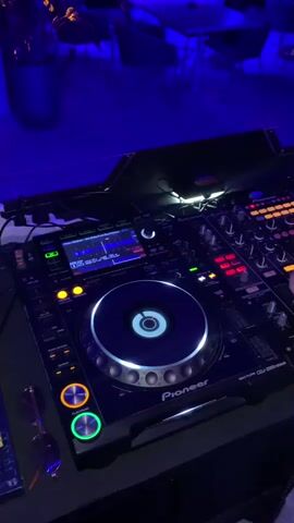 The video shows a close-up view of a DJ setup in a dimly lit room. The primary focus is on a Pioneer DJ controller and mixer. The DJ controller features a large platter with blue and white lighting, along with various buttons and knobs. The mixer has multiple channels with illuminated faders and EQ controls. A person's hand is visible on the mixer, adjusting controls. In the background, blurred tables and chairs are visible under blue and purple ambient lighting, suggesting a nightclub or bar setting. A pair of sunglasses rests on the surface next to the DJ equipment. The video shows the lights on the DJ equipment flashing and changing colors as the music plays.