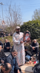 Man in white robe displays framed item to outdoor gathering in Tehran