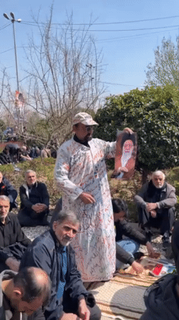 Man in white robe displays framed item to outdoor gathering in Tehran