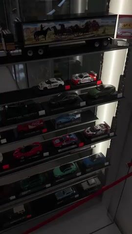 This is a close-up, eye-level, vertical video of a display case filled with model cars. The display case is lit from within, with a bright white light illuminating the shelves and the cars. The shelves are made of black material and are arranged horizontally. The cars are of various colors and styles, and are displayed in individual black stands with labels. In the background, there is a blurred image of a person's leg wearing dark pants, which suggests that the viewer is standing in front of the display case. The video starts with a close-up of the top shelf, showing a large model truck with a Western-themed image on its side. The camera then pans down the display case, revealing more shelves filled with model cars, including sports cars, race cars, and classic cars. The video ends with a shot of the bottom shelf, which is mostly in shadow. The overall impression is that of a well-organized and curated collection of model cars.