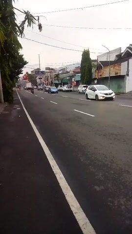 The video shows a street scene with a road that curves to the left and a sidewalk on the left. Several cars are on the road, moving in the same direction. One white car is in the foreground on the right side of the road. A few trees are visible on the left side of the road, and buildings are visible on the right side of the road. The sky is overcast.