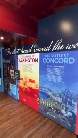 The video captures an indoor exhibition space, likely a museum or historical center, featuring large informational panels about the American Revolutionary War. The floor is made of wood, and the walls are dark red and blue.

Prominently displayed above the panels is a large white text that reads, "The shot heard 'round the world."

The panels visible from left to right include:
- A partially visible panel titled "PAUL REVERE."
- A panel titled "THE BATTLE OF LEXINGTON," depicting a scene of battle.
- A panel titled "THE BATTLE OF CONCORD," with a subtitle "Clash at Concord Bridge," showing soldiers on a bridge. This panel also features a quote with a small portrait.
- A panel titled "THE BATTLE OF BUNKER HILL," illustrating a battle scene and including small portraits and text boxes with quotes.

To the right of the "Bunker Hill" panel, a large framed portrait of George Washington is displayed. He is depicted in colonial attire, wearing a black coat and a white cravat, pointing with his right hand.

Throughout the video, background audio of people speaking can be heard, suggesting other visitors or an audio guide. The camera slowly pans to the right, showing more of the exhibition.