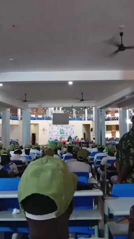 The video shows an indoor gathering of people, likely a conference or an event, in what appears to be a hall or auditorium. The majority of the attendees are wearing green caps and white shirts, suggesting they might be part of a uniform group, possibly youth corps members given the location context of Saakpenwa, Nigeria.

In the foreground, several people in green caps are seated at tables, facing a stage area. The stage has a large banner with text, including "NYSC RIVERS". This confirms the event is related to the National Youth Service Corps in Rivers State, Nigeria. There are speakers or presenters on the stage, and a crowd of attendees is seated in rows of tables and chairs.

A notable detail is a person in a camouflage uniform standing in the aisle on the right side of the frame, observing the proceedings. The ceiling has fans and lights, indicating an indoor setting, likely during the daytime as there is ample artificial lighting. The atmosphere appears to be formal and organized, with a sense of collective participation. The seating arrangement and the presence of a stage with a banner suggest a presentation, lecture, or inauguration ceremony is taking place. The overall scene depicts a structured gathering of young people in Nigeria, likely for official NYSC activities.