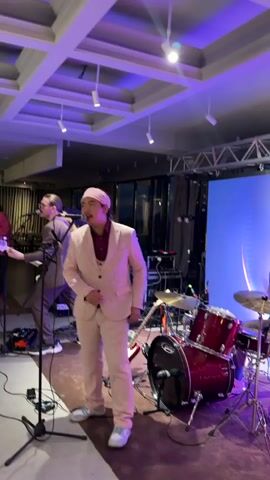 A band is performing on a stage. A lead singer in a pink suit and hat is singing into a microphone while playing a harmonica. A guitarist in a light brown suit is on the left, playing an electric guitar. A drummer in a light brown suit is on the right, playing a red drum set. The stage has purple lighting and a large screen with a blue and white design in the background. There are speakers, cables, and other stage equipment visible. The overall atmosphere appears to be a live music performance in an indoor venue.