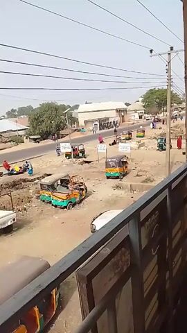 The video shows a street scene in what appears to be a developing country. The view is taken from an elevated position, looking out over a dusty intersection. Several green and yellow auto rickshaws, also known as tuk-tuks, are visible, both parked and in motion. Some people are walking or standing near the rickshaws. A large blue truck passes through the intersection. There is a building in the background. Power lines run across the sky. The video is taken from behind a metal railing.